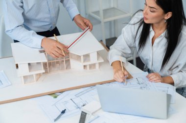 Profesyonel Asyalı erkek mimar ev modelinin uzunluğunu ölçmek için cetvel kullanırken, beyaz tenli genç meslektaşı ev modeliyle toplantı masasındaki verileri analiz etmek için dizüstü bilgisayar kullanıyor. Tertemiz..