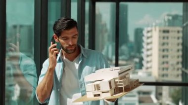 Gülümseyen Kafkasyalı mühendis ev modelini sağa sola çeviriyor ve iş arkadaşına telefon ederken hata noktasını kontrol ediyor. Profesyonel yönetici teftiş, kontrol, mimari modele bakmak. İzleme
