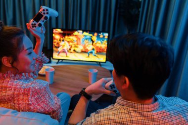 Joystickli birkaç oyuncu dövüş oyunu oynuyor, kadın oyuncu yarışması derecesi arka tarafta TV ekranında modern rahat evinde neon ışıklı oturma odasındaki koltukta oturuyor. İnfobahn.