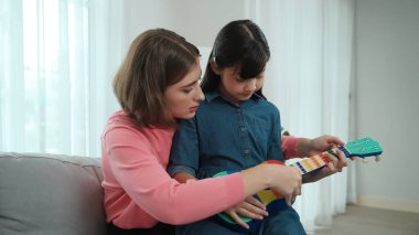 Kafkasyalı bir anne evde akustik müzik öğretirken ukulele çalan mutlu kız. Sevimli çocuk enstrüman öğreniyor. Kafkasyalı mutlu anne ve kız birlikte vakit geçiriyorlar. Pedagoji.