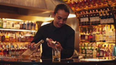 İyi görünümlü Asyalı barmenin içki şurubunu barın lüks içki tezgahında buzlu cama dönüştürdüğü profili. Gece kulübü lüksünde profesyonel barmen kostümü. Vinosity.
