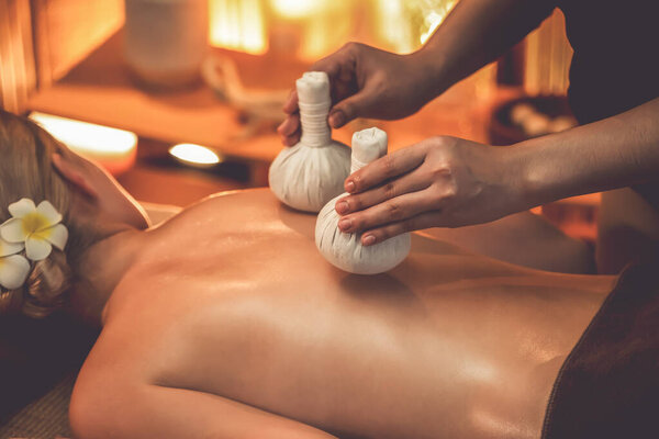 Hot herbal ball spa massage body treatment, masseur gently compresses herb bag on woman body. Tranquil and serenity of aromatherapy recreation in warm lighting of candles at spa salon. Quiescent