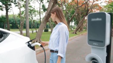 Yavaş çekimde genç bir kadın sonbaharda ulusal parktaki EV şarj istasyonunda EV elektrikli arabaları şarj ediyor. Tatil ve sonbahar sezonunda çevre dostu arabalarla seyahat. Yararlı