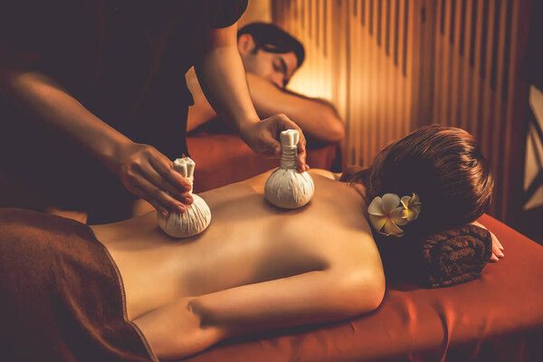 Hot herbal ball spa massage body treatment, masseur gently compresses herb bag on couple customer body. Serenity of aromatherapy recreation in warm lighting of candles at spa salon. Quiescent