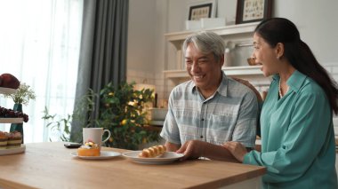 Enerjik büyükanne kahvaltı yapıyor ve dedesi modern mutfakta otururken kocasına servis yapıyor. Gülümseyen yaşlı çift birlikte vakit geçiriyor. Emekli yaşam tarzı konsepti. Myrmidon.