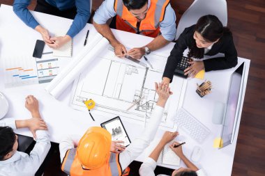 Mimari proje üzerinde birlikte çalışan, inşaat planını gözden geçiren ve toplantı masasında plan oluşturan çeşitli inşaat mühendisi ve müşteri gruplarından oluşan üst düzey bir pankart. Tedbirli
