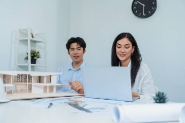 Profesyonel Asyalı erkek mimar ev modelinin uzunluğunu ölçmek için cetvel kullanırken, beyaz tenli genç meslektaşı ev modeliyle toplantı masasındaki verileri analiz etmek için dizüstü bilgisayar kullanıyor. Tertemiz..
