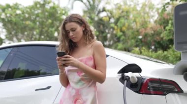 Genç bir kadın doğadaki EV şarj istasyonunda elektrik için akıllı telefon kullanıyor. Modern çevre ve EV araçlı sürdürülebilir otomobil taşıma yaşam tarzı. Titiz