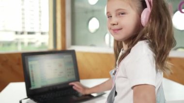 Masaya dizüstü bilgisayarı yerleştirip kameraya el sallayan gülümseyen kız. BTMM sınıfında dizüstü bilgisayar programlama ya da kodlama programı yaparken kulaklık takan bir çocuk gülümsüyor. Etkinlik.