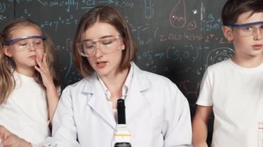 Öğretmen tavsiye verirken beyaz çocuk kimyasal sıvıyı karıştırıyor. Profesyonel eğitmen labaratuar kıyafeti giymiş, renkli solüsyonla dolu deney kabıyla sofrada çeşitli öğrencileri arıyor. Etkinlik.