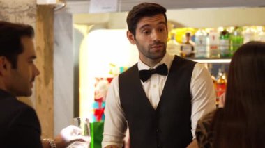 İyi görünümlü barmen hoş geldin içkisi veriyor. İçki tezgahındaki müşterilere dostça sohbetler ediyor. Temiz profesyonel barmen kostümünün içinde harika bir ilk izlenim bırakıyor. Vinosity.