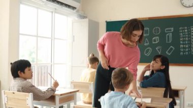 Zeki Asyalı öğrenci, ilkokulda öğretmen yürürken ve ödevlerini kontrol ederken matematik sorusunu sorarak elini kaldırdı. Öğretmen not defterini alırken soruyu cevaplıyor. Pedagoji.