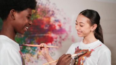 Resim dersinde, renkli duvarda, boyalı beyaz gömlekleri boyayarak gülümseyen farklı çocuklar. Tecrübe alışverişini temsil etmek, birbirimizi öğrenmek. Öğretim