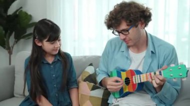 Beyaz tenli çekici baba ukulele çalarken akıllı kız şarkı söylerken onu dinliyor. Yetenekli baba kanepede müzik dinleyip dans ederken gitar çalar. Aile aktivitesi konsepti. Pedagoji.