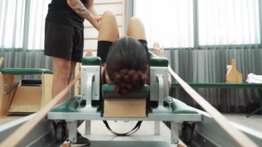 Eğitmen, müşteriye pilates reformatöründe rehberlik ediyor. Vücut geliştirme egzersizinde, vücut hareketlerine, esnekliğine ve beden kuvvetine odaklanmak için kayışlar kullanıyor. Asyalı kız pilates makinesinin önünde yatıyor. Alışkanlık.