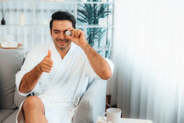 Elinde taze salatalık dilimleri tutan ve bornoz giyen bir adam tatil köyünde ya da otelde yüz bakımının keyfini çıkarıyor. Yüz ve güzellik tedavisi için cilt bakımı. Sessiz.