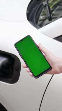 Taklit uzay için şarj istasyonuna bağlı elektrikli aracın yeşil ekran enerji durumunu gösteren bir model telefon. Temiz çevre için ilerici bir kavram. EV, yeşil yenilenebilir enerjiden güç alıyor.