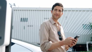 Ağır çekimde çalışan adam, alışveriş merkezinin otoparkındaki kamu EV şarj istasyonunda elektrik için akıllı telefon kullanıyor. EV aracıyla modern çevre ve sürdürülebilir kentsel yaşam tarzı