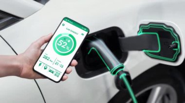 Akıllı telefon akü durumu arayüzünü elinde tutan iş kadını akıllı EV mobil uygulama ile otomobil parkındaki şarj istasyonundan elektrik şarj eden çevre dostu EV araba. İnceleyin