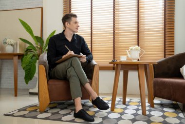 Psikiyatri ofisinde ya da terapi odasında koltukta oturan mutlu ve gülümseyen erkek psikolog portresi. Arkadaş canlısı ve profesyonel ruh sağlığı danışmanı ve terapisti. Açılıyor