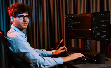 Ekranda yazılım geliştirme bilgisine karşı oturan gülümseyen bilişim geliştiricisinin profili, web sitesindeki son sürüm uygulaması için program kodlaması oluşturarak çevrili kameraya bakıyor. Toplayıcı.