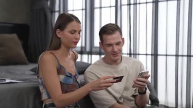Genç çift, kredi kartıyla ödemek için internet üzerinden ödeme uygulaması ve dijital cüzdan kullanarak yatak odasında oturuyor. Mobil internet üzerinden alışveriş ve modern alımlar. Düzenle