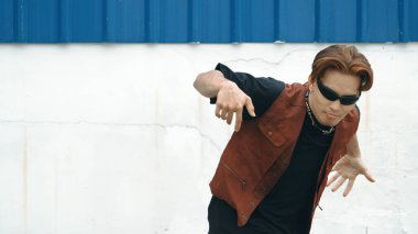 Beyaz arka planda break dans pratiği yapan yakışıklı bir sokak dansçısı. Hip hop tarzı giyinen sporcu, mavi arka planlı bir sokakta müzik eşliğinde ayak sesleri çıkarıyor. Açık hava sporu 2024 hiphop..