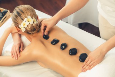 Lüks tatil köyündeki kaplıcalarda sıcak taş masajı. Gün ışığı sükunet ortamında, mutlu kadın müşteri bazalt taş masajından zevk alıyor. Rahatlatıcı bir sıcaklıkla vücudun üzerinde süzülüyor. Sessiz.