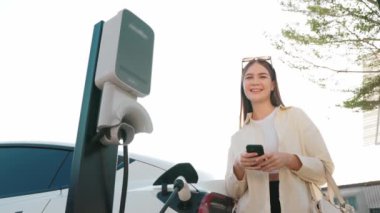 Ağır çekimde genç bir kadın, alışveriş merkezinin otoparkındaki şarj istasyonundan şarj olan EV 'nin pilini şarj ederken akıllı telefondan akü durumunu kontrol ediyor. Modern kadın elektrikli araçla alışverişe gider. Yararlı