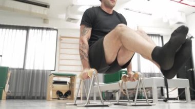 Spor salonunda vücut kontrolünü ve çekirdek gücünü arttırmak için pilates reformcusu üzerinde L-sit egzersizi yapan bir adam. Sportif adam, çekirdek, kol ve omuz güçlendirmek için yenilikçiyi sıkıştırıyor. Alışkanlık.
