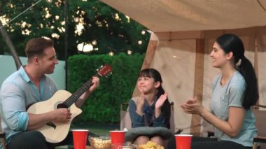 Baba gitar çalarken anne ve kız birlikte kampta oturuyorlar. Ebeveynler, genç nesil çapraz kuşak farklılıklarıyla zaman geçirmek için açık hava kamp aktivitelerini kullanıyorlar. Uyumsuzluk.