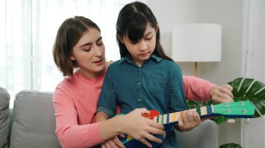 Kafkasyalı bir anne evde akustik müzik öğretirken ukulele çalan mutlu kız. Sevimli çocuk enstrüman öğreniyor. Kafkasyalı mutlu anne ve kız birlikte vakit geçiriyorlar. Pedagoji.