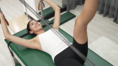 Kadın spor salonunda vücut ve bacakları güçlendirmek için pilates reformatöründe bacak egzersizi yapıyor. Bayan antrenör tek bacak esneme hareketleri yapıyor. Bacak istikrarını arttırmak için Cadillac 'ta kayışlar var. Alışkanlık.