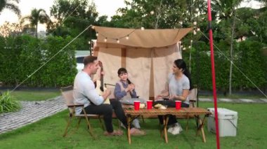 Baba gitar çalarken anne ve kız birlikte kampta oturuyorlar. Ebeveynler, genç nesil çapraz kuşak farklılıklarıyla zaman geçirmek için açık hava kamp aktivitelerini kullanıyorlar. Uyumsuzluk.