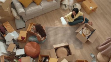 Oturma odasındaki mobilyaların taşınmasına yardım ederken yerde oturan mutlu oda arkadaşı. Yeni eve taşınmak için birlikte çalışan Asyalı lezbiyen aşığının havadan görünüşü. Yer değiştirme konsepti. Myrmidon.