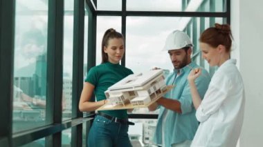 Proje yöneticisi ev modelini elinde tutarken, mimar mühendis takımı da ev tasarımı veya bina inşaatı hakkında fikir alışverişinde bulunuyor. Şehrin penceresinin yanında iç mimar duruyor. İzleme
