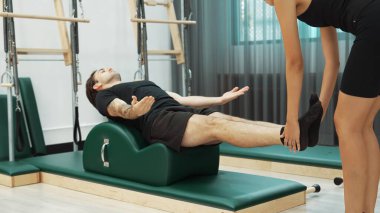 Erkek müşteri pilates ark fıçısında bacak kaldırma alıştırması yapıyor. Vücut dengesini, bacak dayanıklılığını ve spor salonundaki esnekliğini arttırmak için eğitmen yardımı ile birlikte. Öğrenci yalancı ve muziplik pozu veriyor. Alışkanlık.