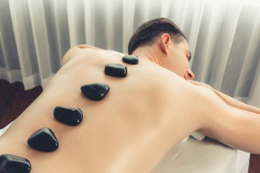 Lüks tatil köyündeki kaplıcada sıcak taş masajı. Gün ışığı dinginliği ortamı, mutlu erkek müşteri bazalt taşı masajının keyfini çıkarıyor. Rahatlatıcı bir sıcaklıkla vücudun üzerinde süzülüyor. Sessiz.