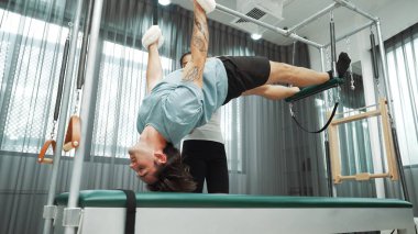 Eğitmen rehberliğinde Pilates Cadillac 'ta ters bacak yükseltme pratiği yapan sağlıklı bir müşteri. Çekirdek dayanıklılığını ve üst vücut dengesini arttırmak için Cadillac 'ı geren adam. Alışkanlık.