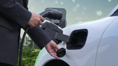 İlerleyen yeşil şehir ESG sembolü, elektrikli araçla birlikte. İş adamı EV arabalarını şarj etmek için şarj aletini şarj ediyor. Şehirdeki şarj istasyonunda ekolojik ve yeşil semboller var..