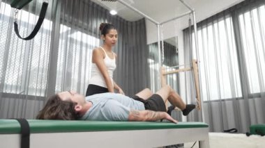 Cadillac ekipmanları üzerinde bacak kaldırma egzersizinde müşteriye yardımcı olan yetenekli pilates eğitmeni. Kadın antrenör, Cadillac 'ın bacak gerdirme hareketine yardım ediyor. Temel bağlantıyı geliştirmek için, bacak esnekliği için. Alışkanlık.