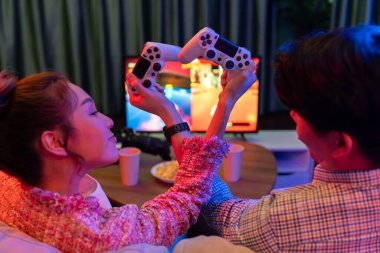 joystickli elleri çapraz oynayan bir çift arka tarafta TV ekranında dövüş oyunu oynuyor, modern rahat evinde neon ışıklı oturma odasında kanepesinde oturuyor. İnfobahn.