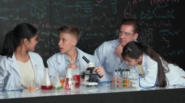 Çok kültürlü bir öğrenci mikroskop altında incelerken güzel bir kız da numuneyi inceliyor. BTMM sınıfında karatahta bilim teorisi deneyi yapan liseli çocuklara bakan öğretmen. Öğretim
