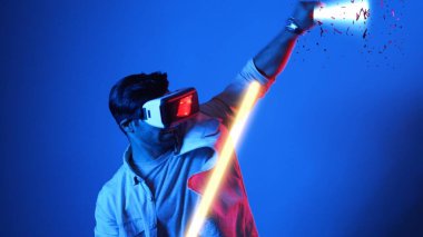 Parlayan kılıcı tutan ve VR gözlük takan oyuncu oyun oynuyor. Çekici erkek model sanal gerçeklik gözlüğü kullanarak yüzen bloğu eskrirken heyecanlanıyor. Teknoloji yeniliği. Aygıt.