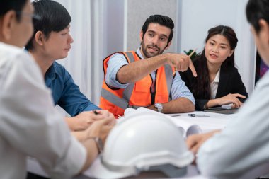 Mimari proje üzerinde birlikte çalışan çeşitli inşaat mühendisi ve müvekkil grupları, inşaat planını gözden geçirip toplantı masasında plan oluşturuyorlar. Tedbirli
