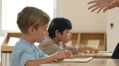 Beyaz tenli ilkokul öğrencisinin tahtada otururken bir nota bakması ve soruya cevap vermesi. Çocuklar derse katılırken genç öğretmen sınıf çalışmasını ya da sınavı açıklar. Pedagoji.