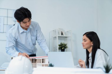 Profesyonel Asyalı erkek mimar ev modelinin uzunluğunu ölçmek için cetvel kullanırken, beyaz tenli genç meslektaşı ev modeliyle toplantı masasındaki verileri analiz etmek için dizüstü bilgisayar kullanıyor. Tertemiz..