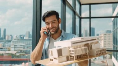 Gülümseyen Kafkasyalı mühendis ev modelini sağa sola çeviriyor ve iş arkadaşına telefon ederken hata noktasını kontrol ediyor. Profesyonel yönetici teftiş, kontrol, mimari modele bakmak. İzleme