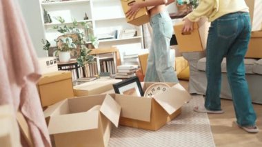 Kutu taşıyan ve kutularla çevrili bir şekilde oturan ve kutuları açmaya hazırlanan çift. Mutlu kız ve arkadaş yeni eve taşınırken kutuları tutuyor ve sohbet ediyorlar. Yer değiştirme konsepti. Myrmidon.