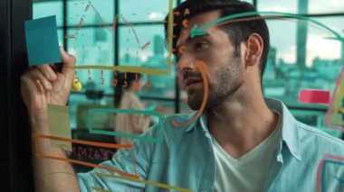 Akıllı proje yöneticisi cam duvara yapışkan notlar ve planlama stratejisi için zihin haritası yazarken, aynı zamanda çok kültürlü iş adamları da şehir manzaralı iş planlarını tartışıyorlar. İzleme